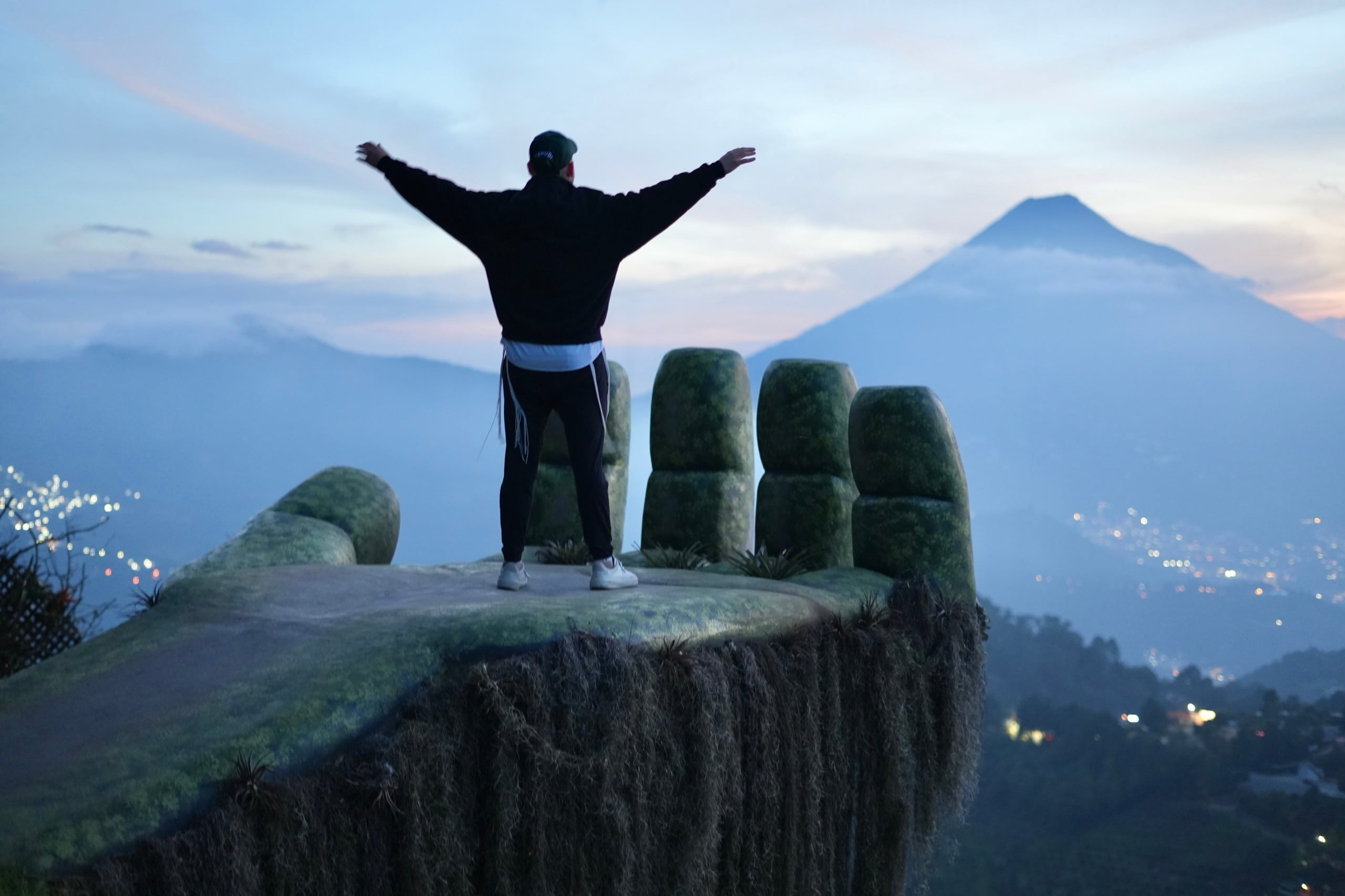Arms wide on the giant hand lookout, volcano and city lights at dusk